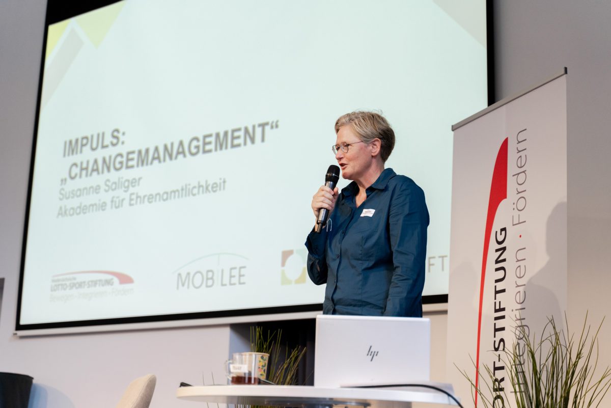 Person steht auf der Bühne und spricht in ein Mikrofon, im Hintergrund steht auf einer Präsentation "Changemanagement".