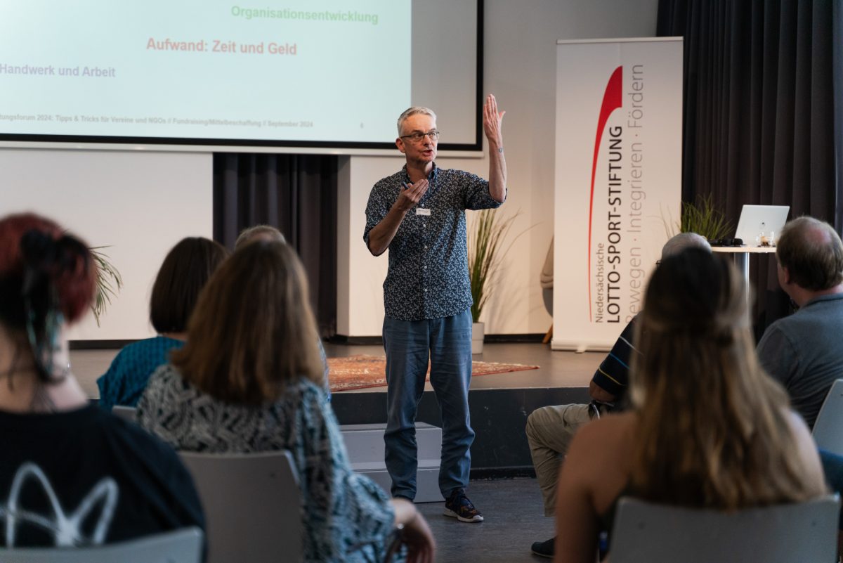 Person steht vor einer Gruppe sitzender Menschen und spricht in ein Mikrofon.