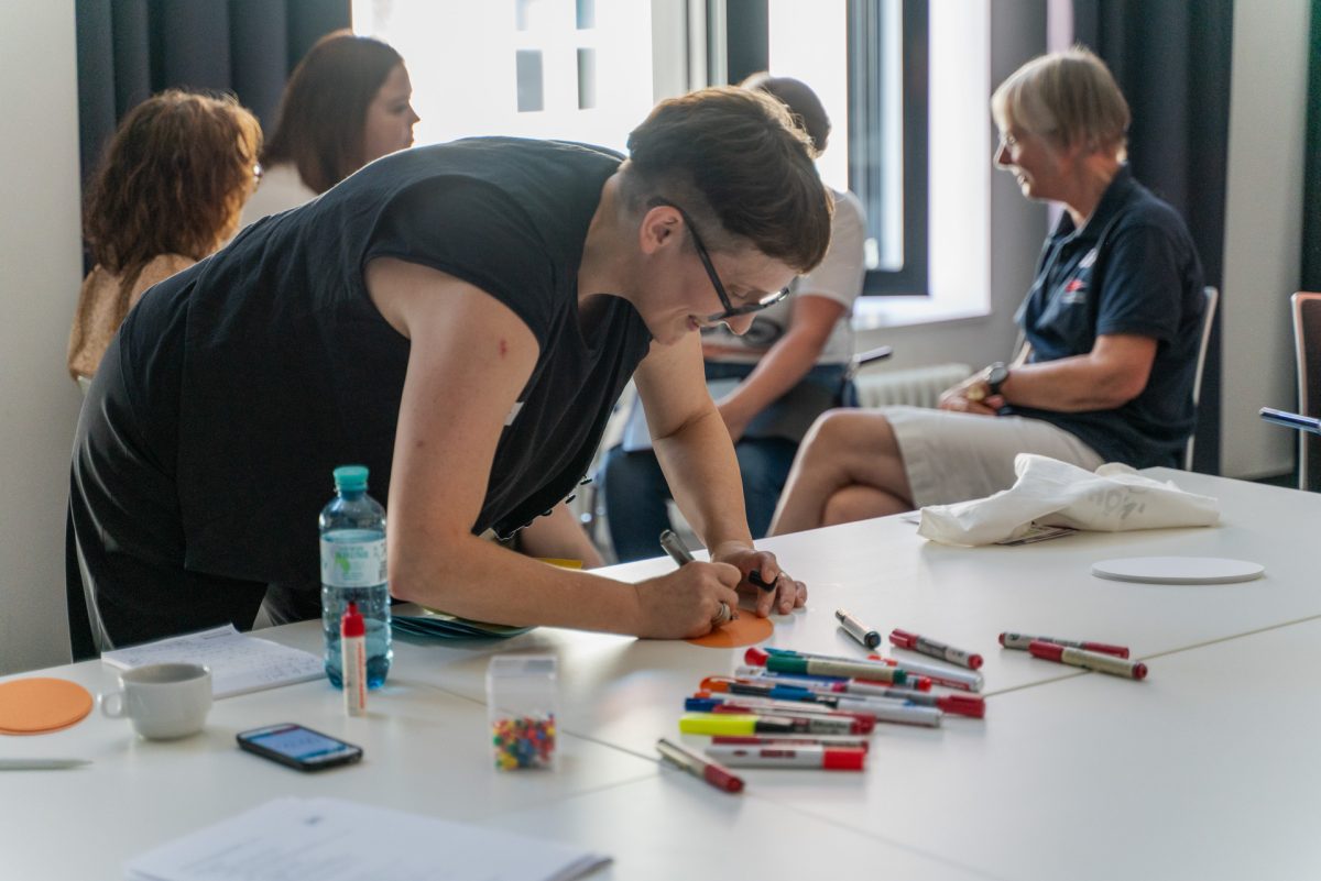 Person steht an einem Tisch voller Stifte und schreibt etwas auf ein Papier.