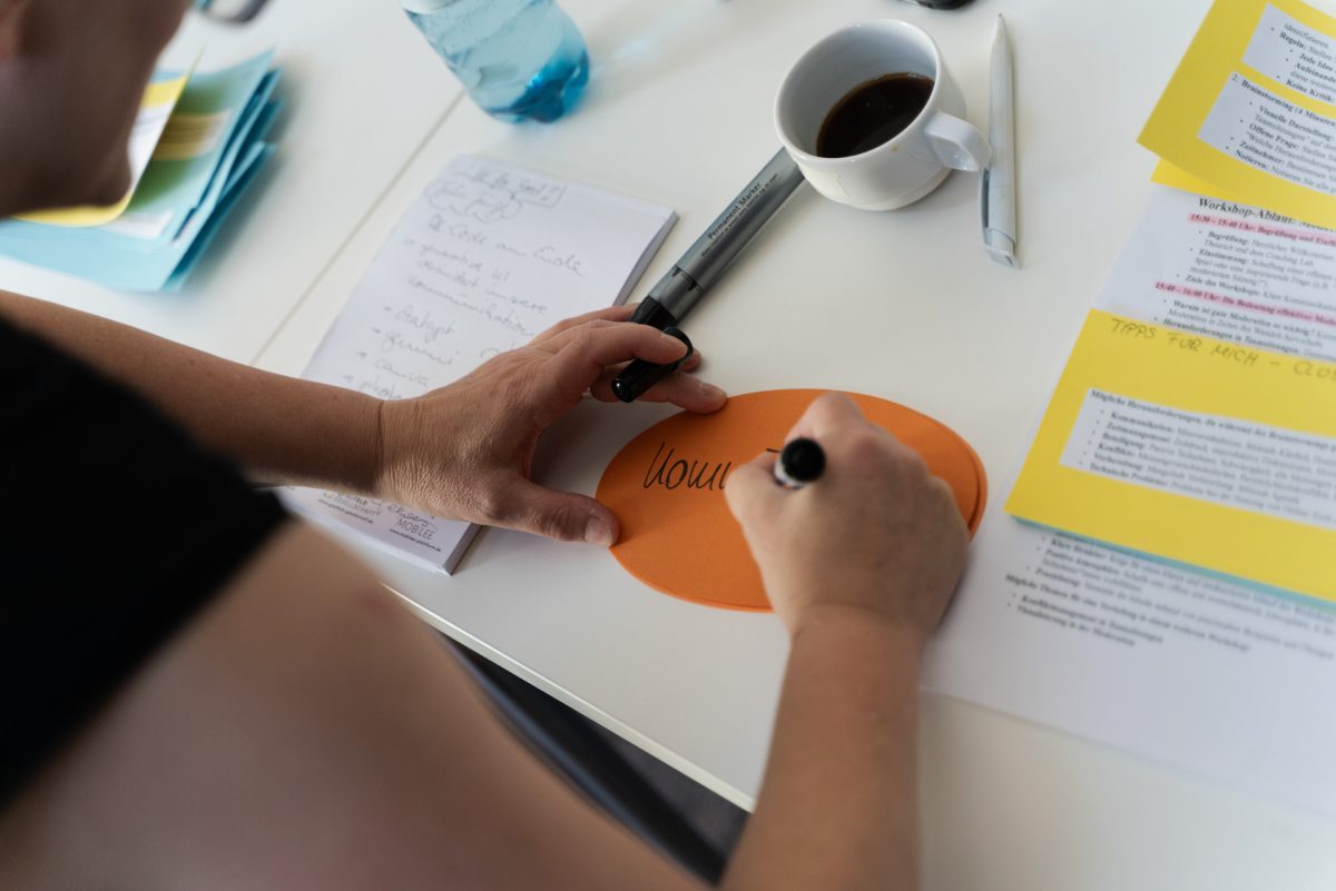 Person schreibt auf einem Kärtchen, daneben liegen Stifte, bedruckte Papiere und ein Becher mit Kaffee.