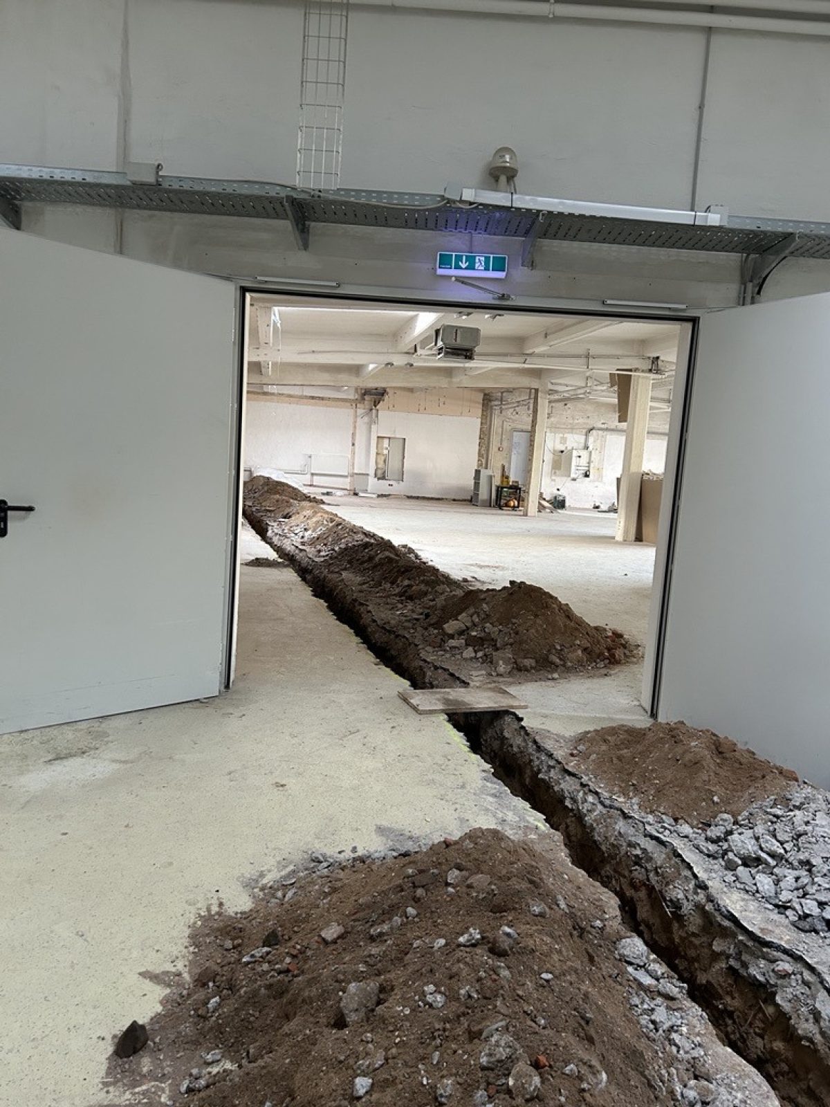 Auf dem Foto ist ein Teil einer großen Halle mit geöffneter Tür zum nächsten Gebäudeteil zu erkennen. Der Boden ist aufgerissen, um dort Rohre neu zu legen.