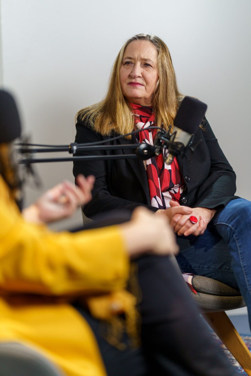 Eine Person sitzt auf einem Stuhl gegenüber einer weiteren Person, die unscharf zu sehen ist. Vor beiden ist ein Mikrofon zu sehen.