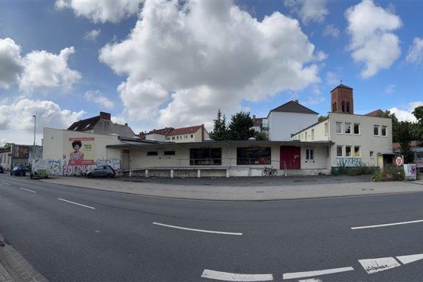 Auf dem Bild ist ein Gebäude zu sehen, welches an einer Straße liegt. Es hat eine große Frontscheibe sowie eine rote Eingangstür. Das Gebäude liegt vor einem Wohngebiet und der rechte Teil ist zweisstöckig.