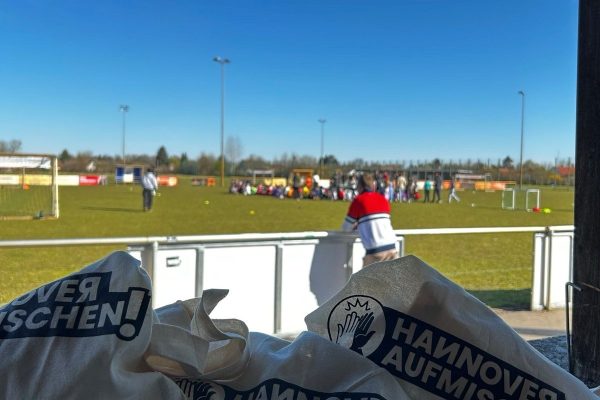 Das Bild zeigt den Blick von einer Tribüne auf einen Sportplatz. Im Vordergrund liegen Jutebeutel mit dem Aufdruck "Hannover aufmischen"