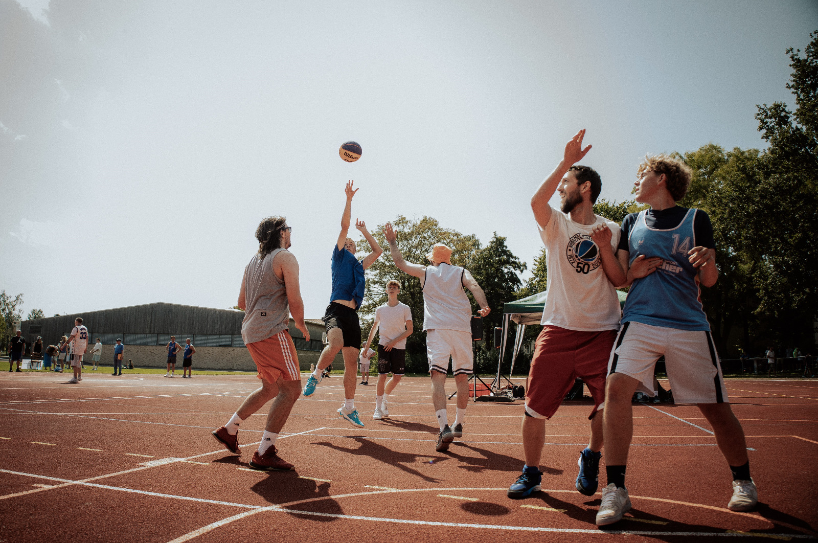 Mehrere junge Männer spielen bei sonnigem Wetter Basketball auf einem Platz im Freien.