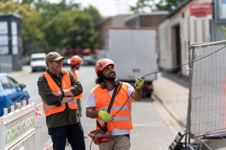 Zwei Männer in orangefarbenen Warnwesten schauen auf einer Baustelle nach oben. Einer trägt einen Schutzhelm und hält ein Kabel in den Händen.