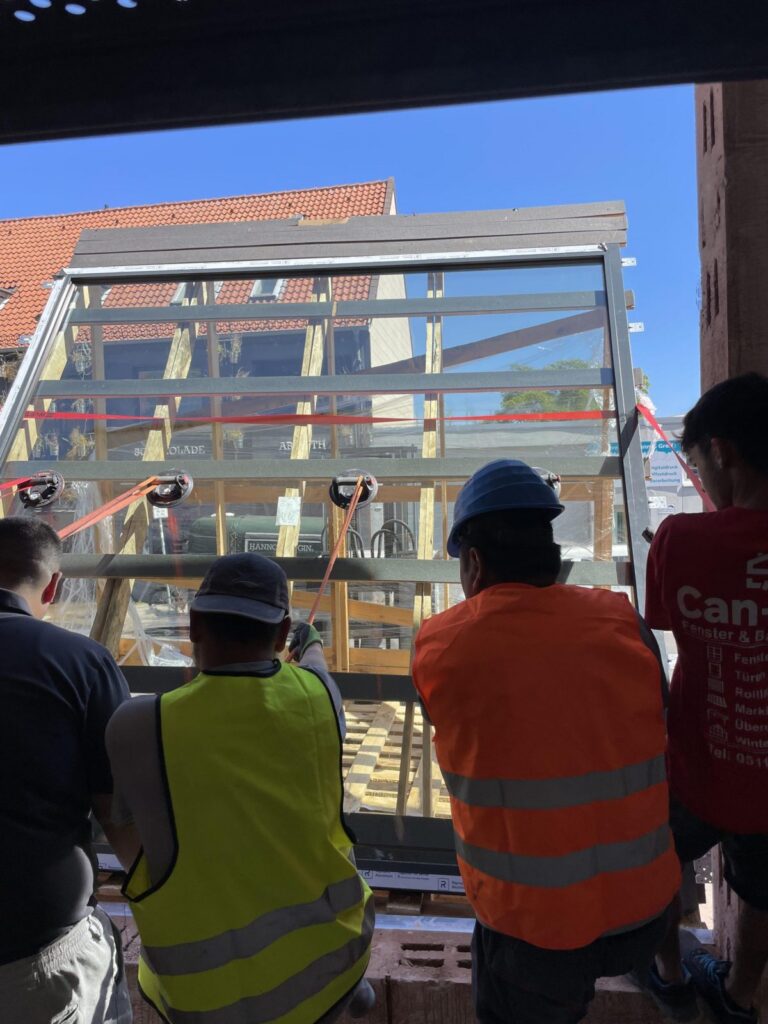 Vier Handwerker setzen gemeinsam ein großes Fensterelement in eine Maueröffnung ein.
