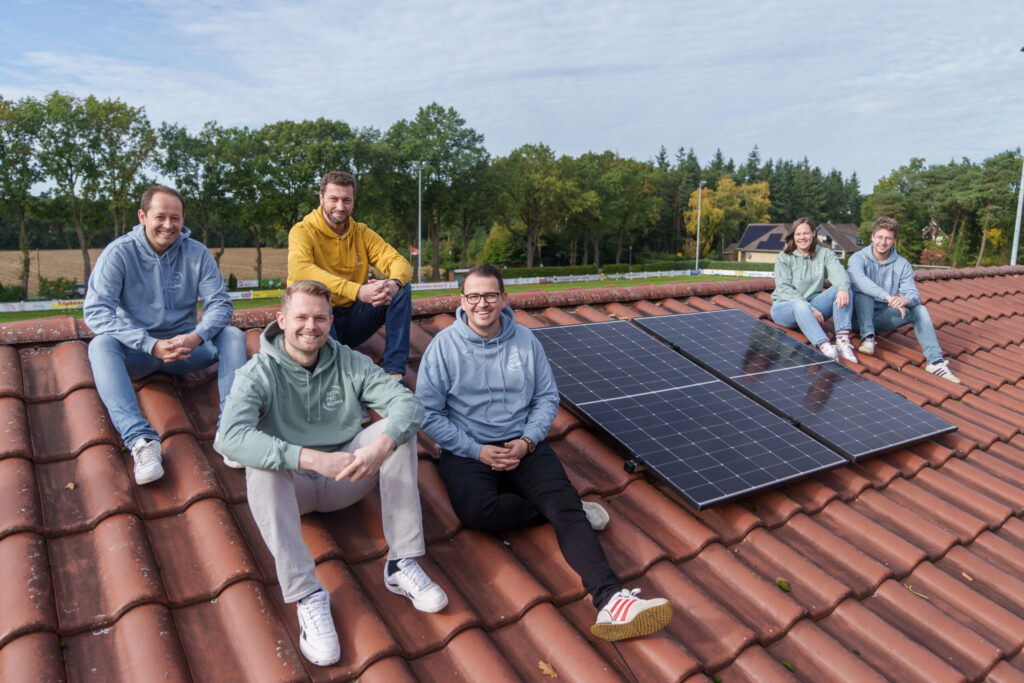Gruppe von sechs Engagierten eines Sportvereins in bunten Hoodies sitzt auf dem Dach des Vereinsheims direkt neben zwei installierten Solarmodulen.