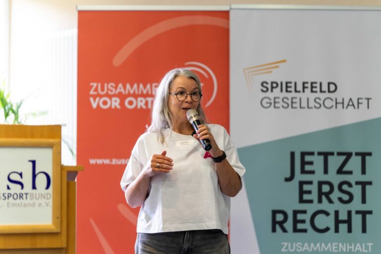 Eine Frau mit Brille spricht in ein Handmikrofon und gestikuliert vor Veranstaltungsbannern.