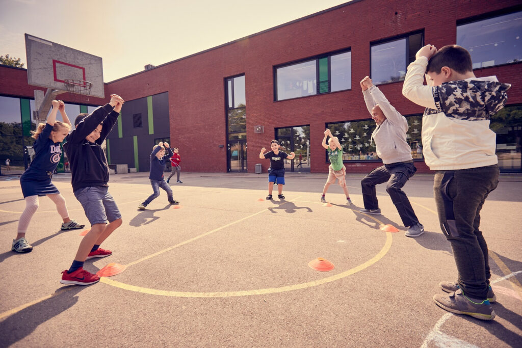 Kinder stehen im Kreis auf einem Schulhof und heben die Arme über den Kopf.