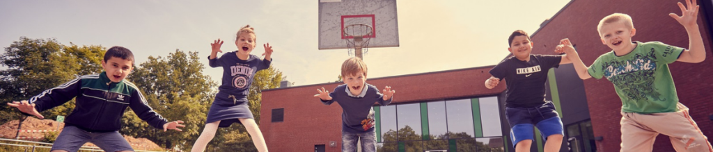 Fünf Kinder springen vor einem Basketballkorb auf einem Schulhof und schauen in die Kamera