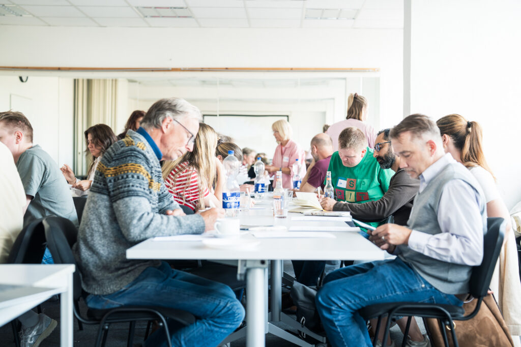 Gruppe von Menschen sitzt an langen Tischen und arbeitet oder liest in einem hellen Raum.