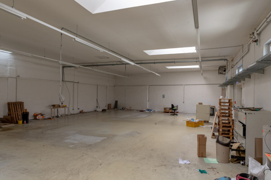 Helle, weitgehend leere Lagerhalle mit Oberlichtern, Paletten, Kartons und einzelnen Möbeln.