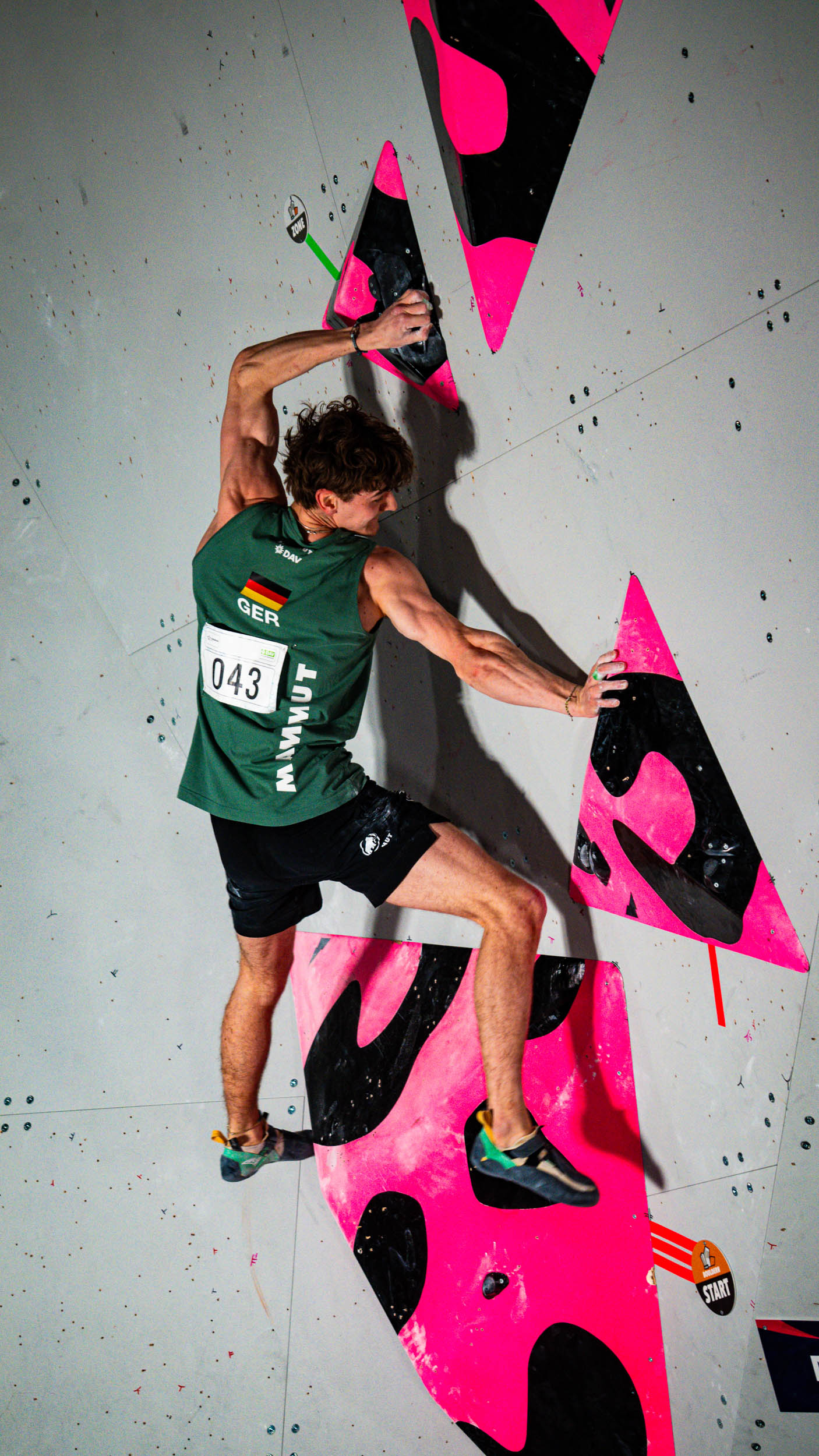 Sportkletterer in grünem Trikot mit Startnummer 043 an einer Boulderwand mit pinken und schwarzen Griffen.