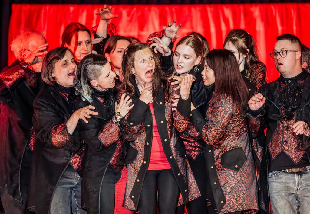 Theatergruppe in dunkelroten Kostümen: Eine Schauspielerin schreit erschrocken auf, die anderen bedrängen sie.