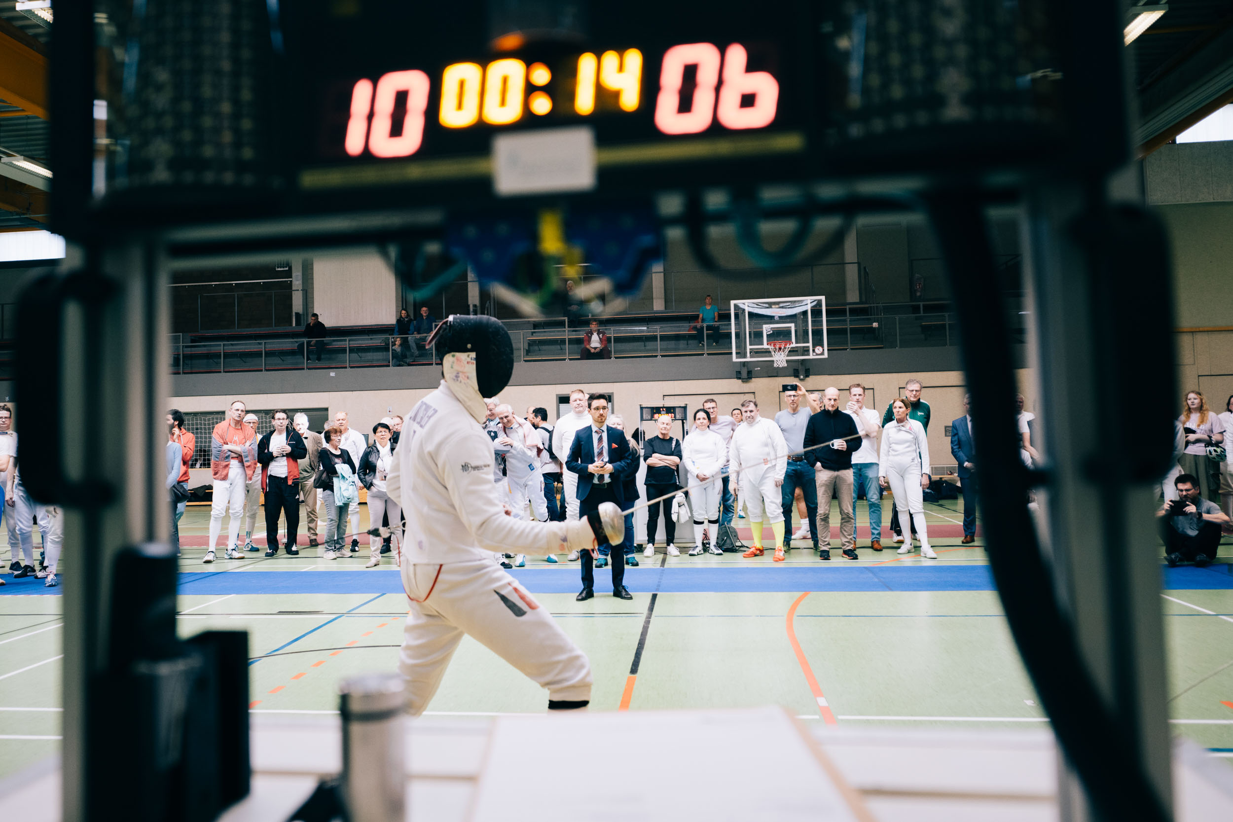 Blick durch eine digitale Anzeigetafel auf ein Fechtturnier in einer Sporthalle: Ein Fechter in weißer Schutzkleidung und Maske tritt vor Publikum und einem Schiedsrichter auf der Fechtbahn an.