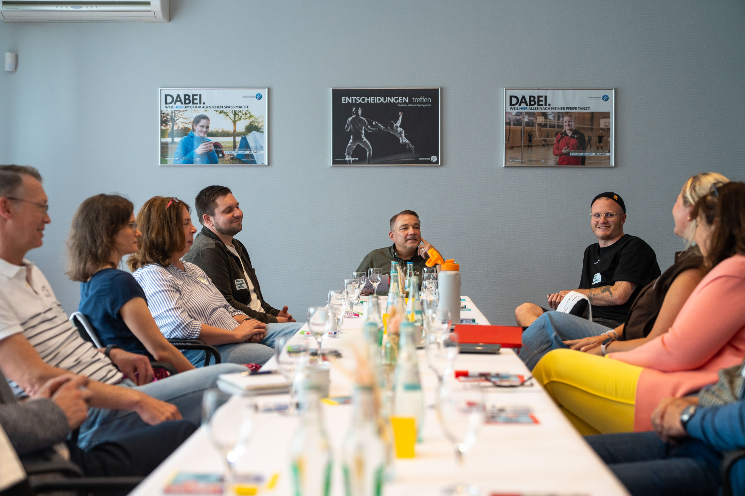 Gruppe von Menschen sitzt an einem langen Tisch in einem Besprechungsraum mit drei Plakaten an der Wand
