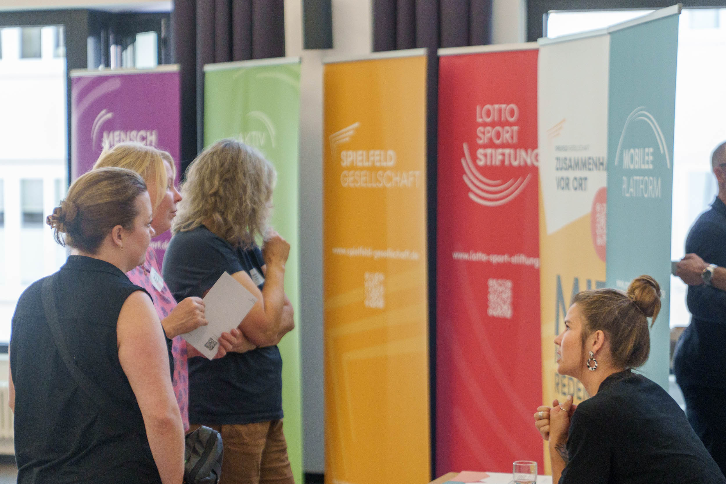 Teilnehmende im Gespräch bei einer Informationsveranstaltung vor bunten Roll-up-Bannern, darunter das rote Banner der Lotto-Sport-Stiftung und weiteren Projekten.
