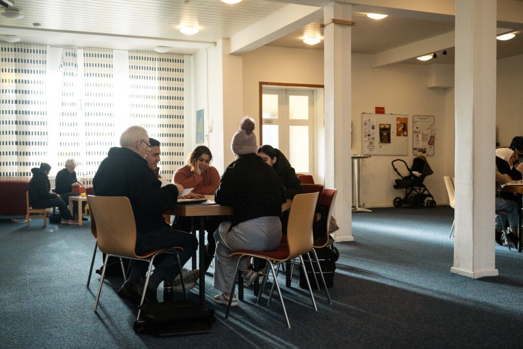 Eine kleine Gruppe von Menschen unterschiedlichen Alters sitzt zusammen an einem Tisch in einem Gemeinschaftsraum und bespricht Unterlagen.