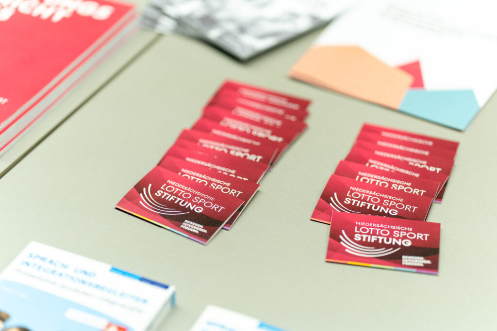 Verschiedene Informationsmaterialien und Flyer auf einem Tisch, darunter mehrere Stapel roter Visitenkarten der Niedersächsischen Lotto-Sport-Stiftung mit weißem Logo und Schriftzug.