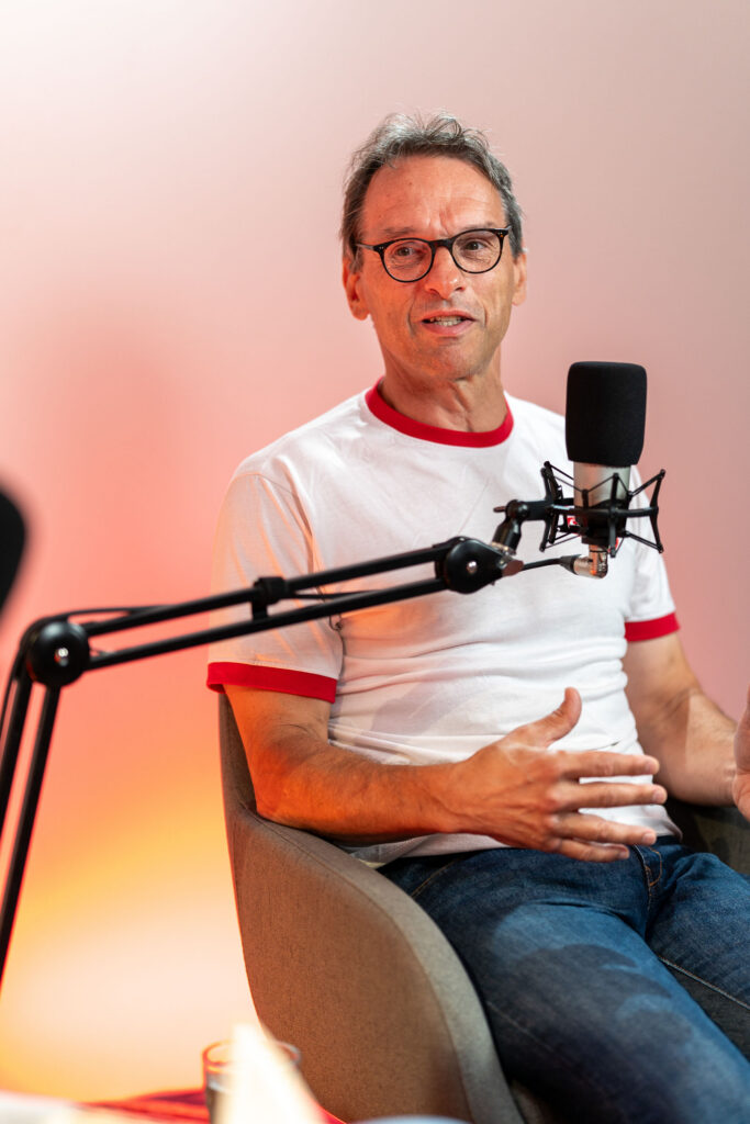 Mann mit Brille und weißem T-Shirt mit rotem Kragen sitzt in einem Sessel vor einem Mikrofonarm und spricht