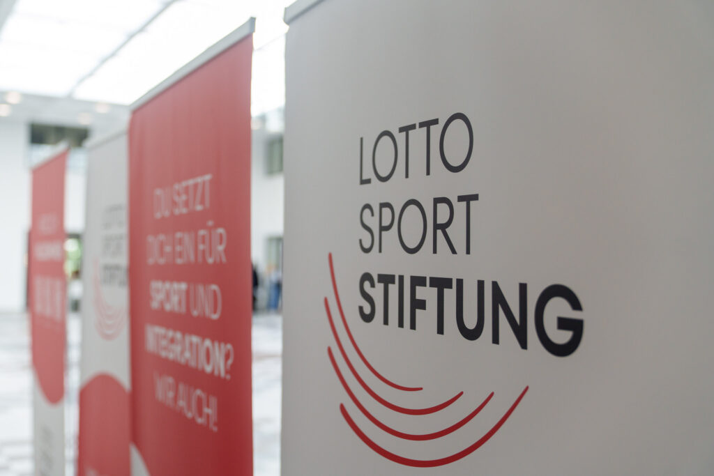 Mehrere aufgestellte Roll-up-Banner bei einer Veranstaltung; im Vordergrund das weiße Banner der Lotto-Sport-Stiftung mit Firmenlogo und Schriftzug, im Hintergrund rote Infobanner zu Sport und Integration.
