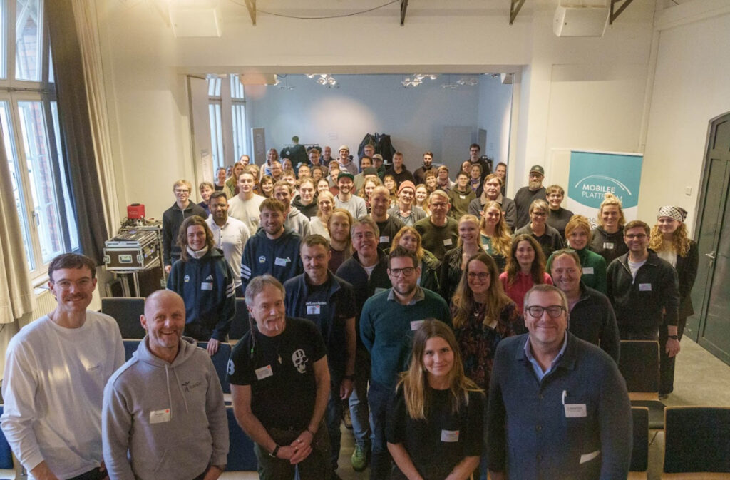 Gruppe von Menschen in einem hellen Raum mit großen Fenstern, die in die Kamera lächeln