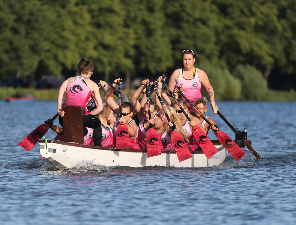 Ein Frauen-Drachenboot-Team in rosa Trikots paddelt mit roten Paddeln im Gleichschritt über ein Gewässer, während eine Steuerfrau im Stehen das Boot am Heck lenkt.