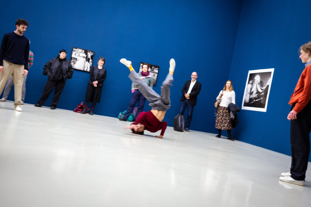 Ein Tänzer führt im Zentrum eines Museumsraums vor kornblumenblauen Wänden einen Einhand-Handstand (Breakdance-Freeze) aus, während umstehende Besucher fasziniert zuschauen.