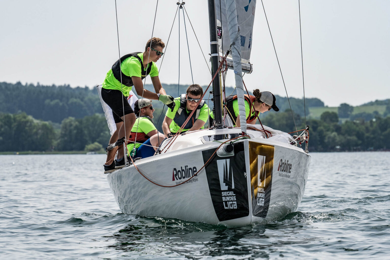 Vier Personen in leuchtenden Trikots und Schwimmwesten bedienen gemeinsam ein Segelboot auf einem See.