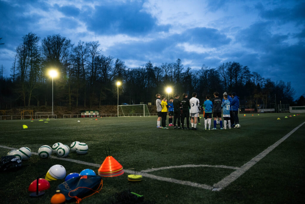 Eine Jugend-Fußballmannschaft versammelt sich bei Dämmerung unter Flutlicht zu einer Besprechung auf dem Kunstrasenplatz, während im Vordergrund Trainingsutensilien wie Fußbälle und Markierungshütchen liegen.